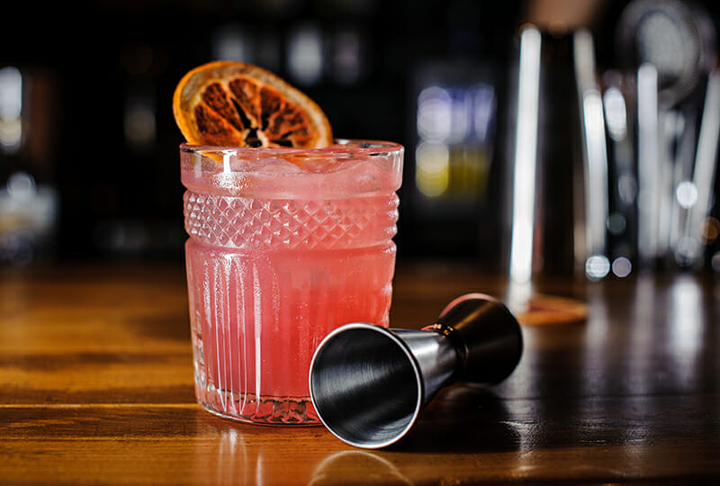 Glass of pink alcoholic cocktail decorated with a slice of orange on the table against the indoors background of a bar