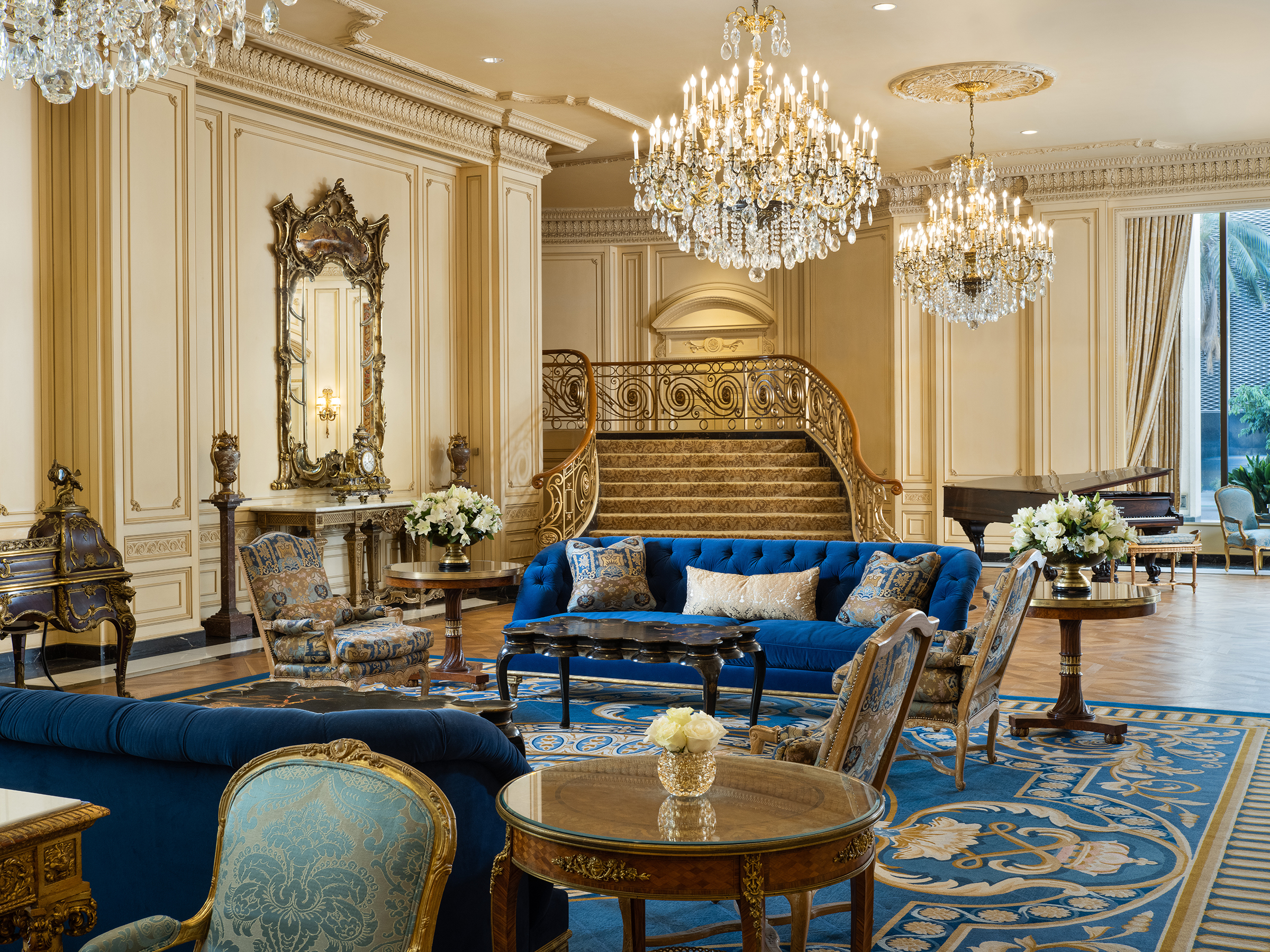 A luxurious hotel lobby with a staircase, chandeliers, and comfortable seating.